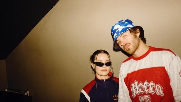 Ein junges Pop-Duo, sie mit dunklen Sonnenbrillen, er mit blau-weißem Freizeithut, blickt in einem Raum mit schrägem Lichteinfall etwas misstrauisch in die Kamera.