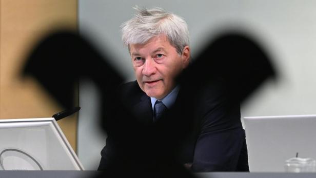Ein älterer Mann im Anzug sitzt an einem Schreibtisch, im Vordergrund ist ein schwarzes Symbol unscharf zu sehen.