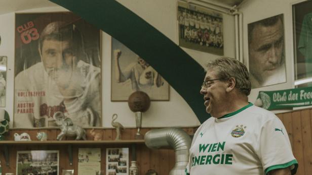 Fanclub-Präsident Martin Reiser in Rapid-Dress steht in seinem Vereinslokal, an der Wand etliche Poster und Fanschals