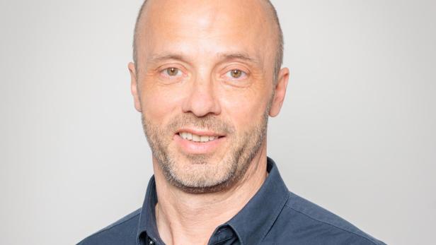 Mann mit kurzem Haar und Bart trägt ein dunkelblaues Hemd und lächelt vor neutralem Hintergrund.