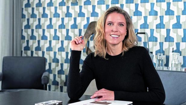 Eine Frau mit blondem Haar, Sigrid Eckhardt, Geschäftsführerin Austropapier, sitzt lächelnd an einem Tisch mit einem Notizbuch und einem Smartphone vor einer gemusterten Wand.