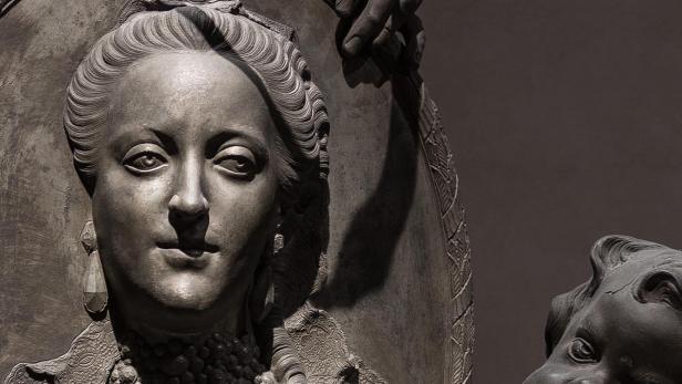 Detail einer Skulptur mit einer Figur in aufwändiger Kleidung und Perlenketten vor einem Schild, darüber eine Schlange und eine Hand