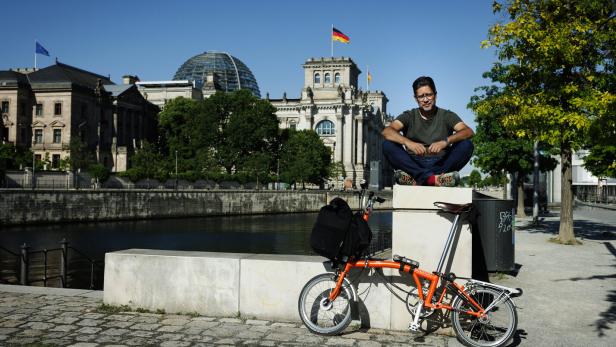 Der Autor Hasnain Kazim sitzt auf einem Mauervorsprung, im Hintergrund ist der Berliner Reichstag zu sehen, im Vordergrund steht ein Falt-Rad