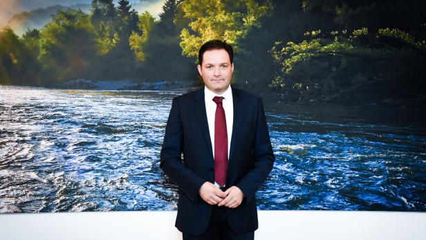 Ein Mann im Anzug mit roter Krawatte, Klimaminister Totschnig, steht vor einem großen Wandbild mit Fluss und bewaldeter Landschaft.