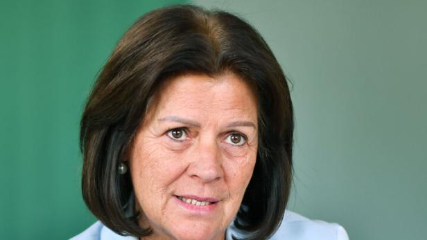 Frau mit dunklen Haaren, hellblauem Blazer und Perlenkette sitzt an einem Tisch und spricht.