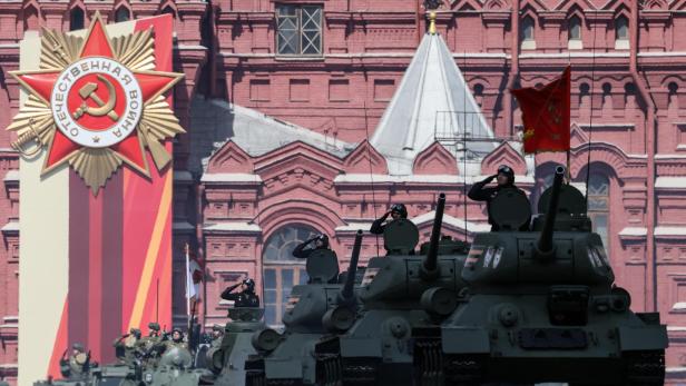 Panzer fahren am Roten Platz, links hinter ihnen ein Emblem einer Hammer und Sichel auf rotem Stern.
