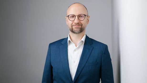 Mann mit Brille und Bart im blauen Anzug vor grauem Hintergrund.