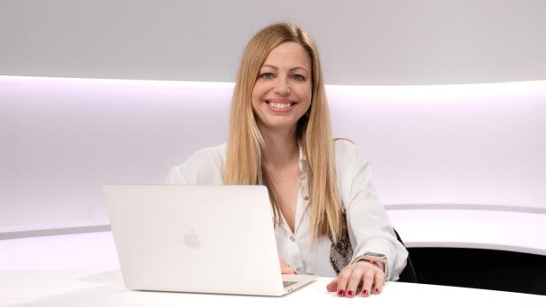 Frau mit langen blonden Haaren lächelt an einem Schreibtisch mit Laptop vor hellem Hintergrund.