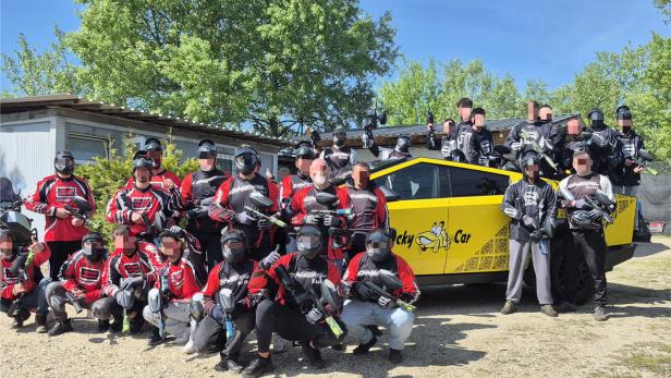 Gruppe von Paintballspielern in Ausrüstung vor einem gelben Auto im Freien bei sonnigem Wetter.