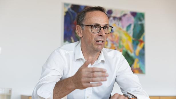 Ein Mann mit Brille und weißem Hemd spricht gestikulierend an einem Tisch, im Hintergrund hängt ein buntes Gemälde.