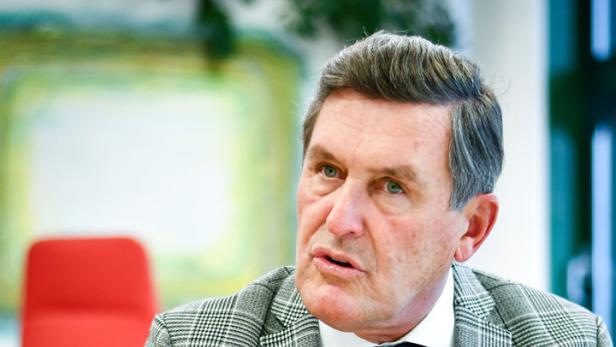 Ein Mann im karierten Anzug sitzt an einem Tisch und spricht mit gefalteten Händen.