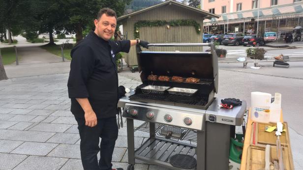 Ein Mann grillt Fleisch auf einem Gasgrill im Freien vor einem Holzhaus und geparkten Autos.