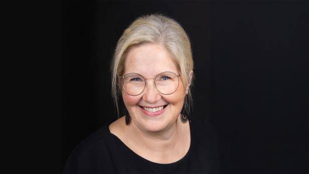 Frau mit blondem Haar, Brille und schwarzem Oberteil lächelt vor schwarzem Hintergrund.
