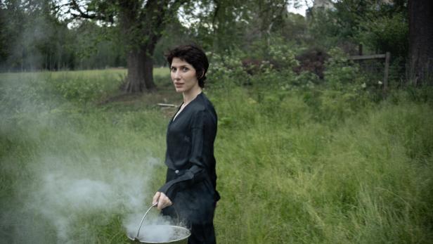 Die Musikerin Aldous Harding trägt einen Kübel, aus dem Dampf strömt, durch eine grüne Waldlandschaft.