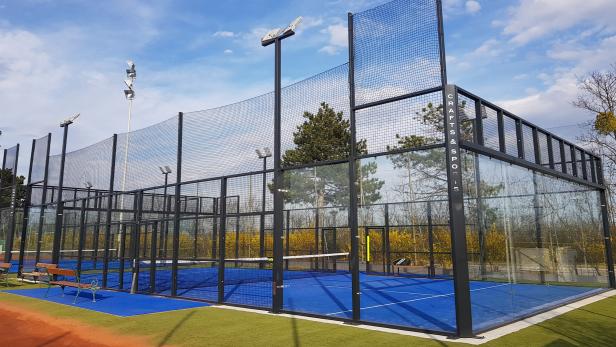 Ein moderner Padelplatz im Freien mit blauen Spielfeldern und hohen Glas- und Gitterwänden unter blauem Himmel.