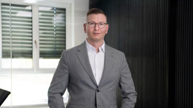 Ein Mann mit Brille und kurzem Haar trägt einen grauen Anzug und steht in einem modernen Büro mit Fenster im Hintergrund.