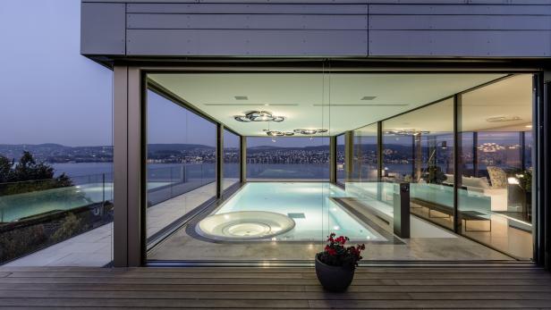Modernes Wohnzimmer mit großem Innenpool, Whirlpool und Panoramablick auf einen See und eine Stadt im Hintergrund.