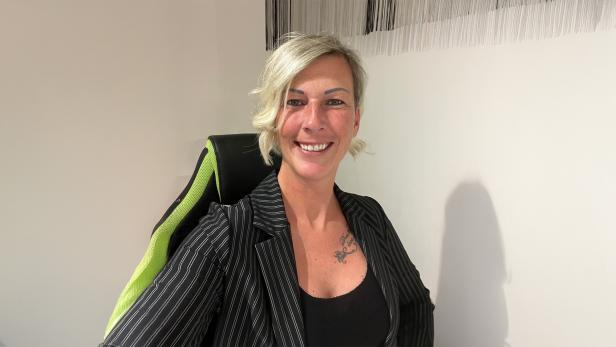 Eine lächelnde Person mit blondem, kurzem Haar sitzt auf einem Stuhl mit grünem Rückenlehnenbezug vor einer hellen Wand.