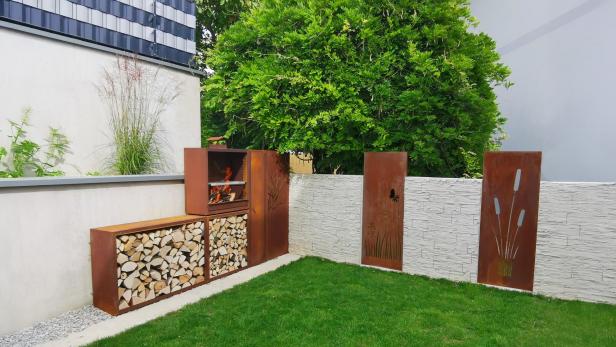 Ein Grill mit Holzlager aus rostigem Metall steht in einer modernen Gartenecke mit grüner Rasenfläche.