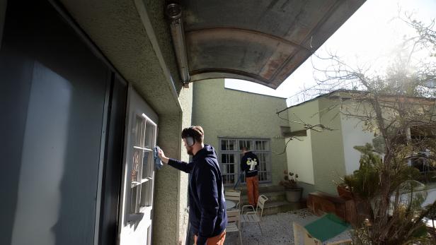 Zwei Personen reinigen Fenster in einem sonnigen Innenhof mit Gartenmöbeln und kahlen Ästen.