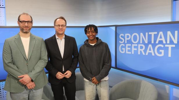 Drei Personen stehen vor einem Bildschirm mit der Aufschrift 'SPONTAN GEFRAGT' in einem Fernsehstudio