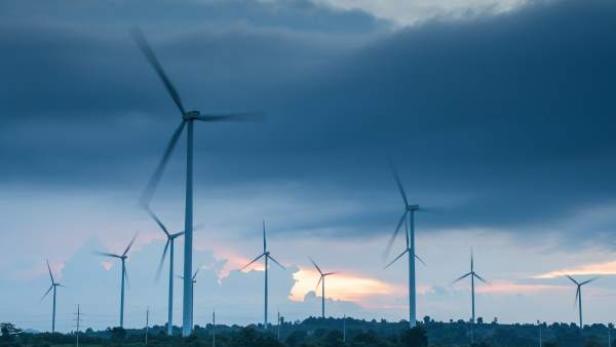 Mehrere Windräder stehen auf einer grünen Landschaft unter einem bewölkten Himmel mit einem hellen Lichtschein am Horizont