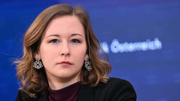 Eine Frau mit Ohrringen und dunklem Blazer blickt ernst in die Kamera vor blauem Hintergrund.