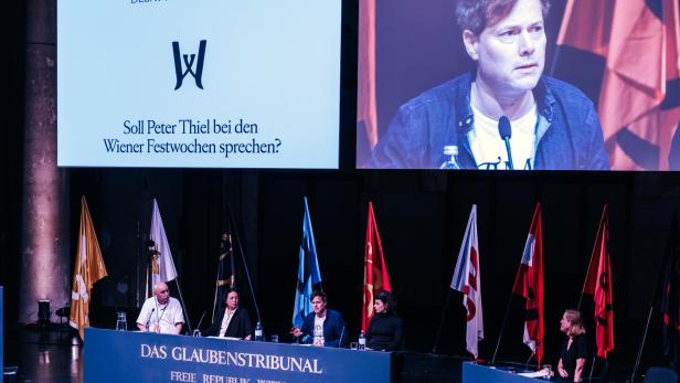 Foto eines beflaggten, mit fünf Menschen besetzten Podiums, auf einem Videoscreen dahinter sieht man Festwochenchef Milo Rau in Großaufnahme.