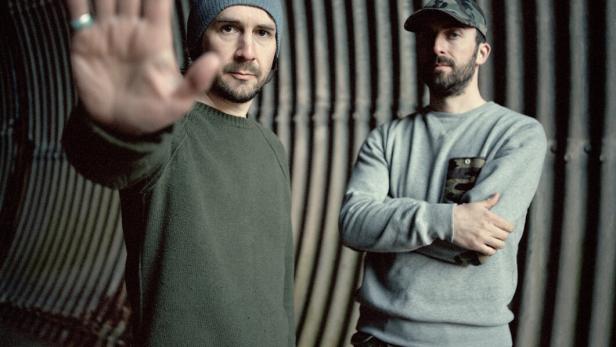 Zwei ernst dreinblickende Musiker in einem kargen Innenraum, der eine mit der verschränkten Armen, der andere hält die offene Hand in die Kamera, als wollte er das Foto untersagen. 