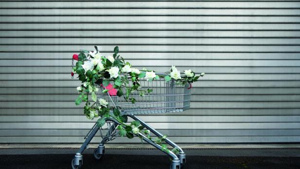 Einkaufswagen mit weißen Blumen und grünem Blattwerk vor einer grauen Metalljalousie