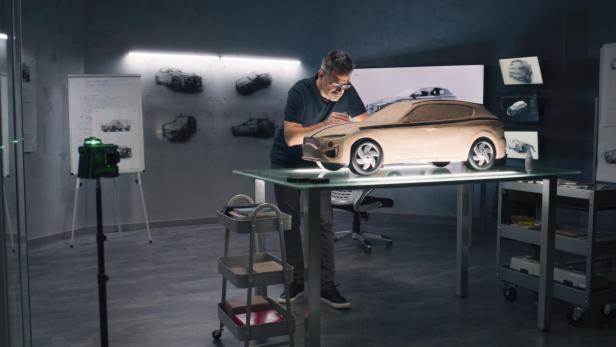 Person steht an einem Tisch und arbeitet an einem braunen Automodell aus Ton in einem Raum mit mehreren Autodesign-Skizzen an den Wänden