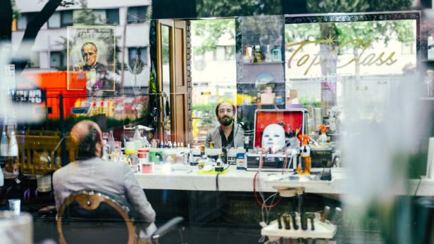 Ein Mann sitzt vor einem Spiegel im Friseursalon „Top Class Hairstyle“, während sich sein Gesicht in der Scheibe spiegelt.