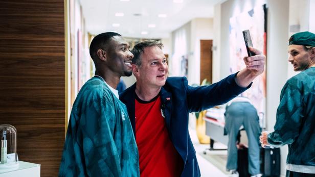 David Alaba lächelt, während ein Mann in einem roten Trikot und blauem Sakko, Sportminister Babler, ein Selfie mit ihm in einem Flur macht.