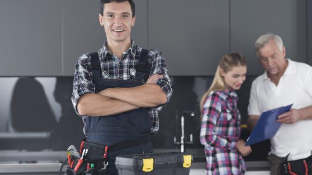 Ein Handwerker steht mit verschränkten Armen und Werkzeugtasche im Vordergrund, während im Hintergrund zwei Personen ein Angebot besprechen.