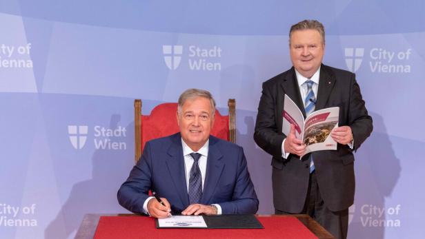 Ein Mann sitzt an einem Tisch und unterschreibt, daneben steht ein zweiter Mann mit einer Broschüre vor Wiener Stadtlogo-Hintergrund.