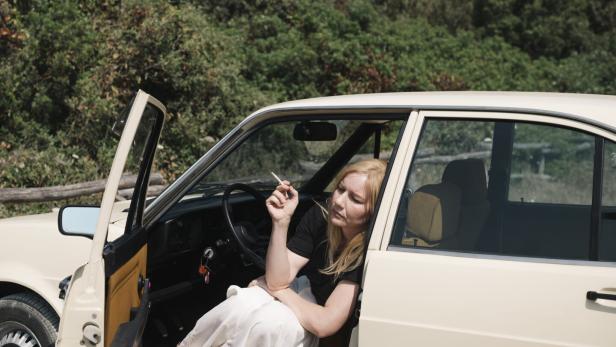 Die Schauspielerin Sandra Hüller mit blonder Perücke, rauchend, in einem geparkten Auto sitzend, im Hintergrund ein Wald
