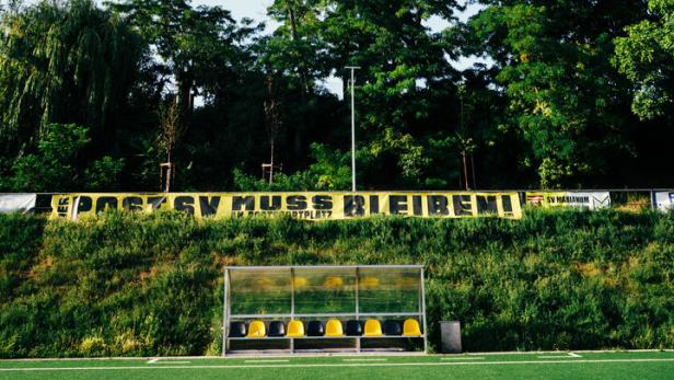 Eine Reihe von Sitzplätzen am Rand eines Fußballplatzes, dahinter Bäume und ein Banner, auf dem steht: "PostSV muss bleiben!"