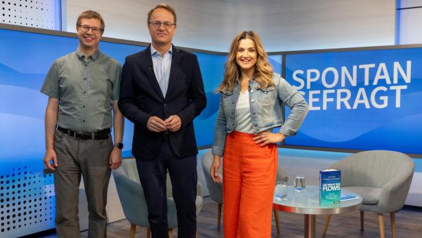 Drei Personen stehen vor einem blauen Hintergrund mit der Aufschrift 'SPONTAN GEFRAGT', links und rechts je ein grauer Sessel und ein runder Glastisch mit zwei Gläsern und einem Buch