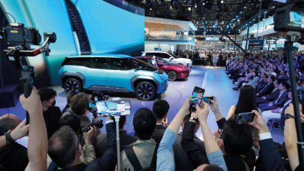 Menschen fotografieren auf einer Messe zwei ausgestellte Autos vor einer großen, gebogenen Bildschirmwand