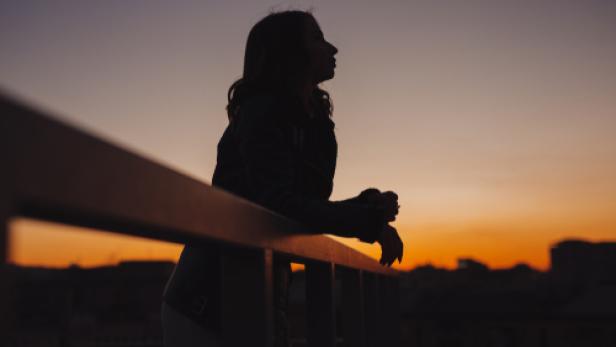 Silhouette einer Person, die an einem Geländer lehnt vor einem orangefarbenen Sonnenuntergangshimmel