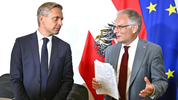Zwei Männer im Anzug, die Minister Hattmannsdorfer und Marterbauer, diskutieren vor österreichischer und EU-Flagge, einer hält Unterlagen in der Hand.