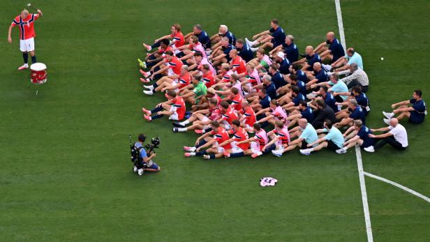 Die Norwegische Nationalmannschaft sitzt auf dem Fußballfeld und macht Ruderbewegungen zum Trommeltakt von E. Haaland