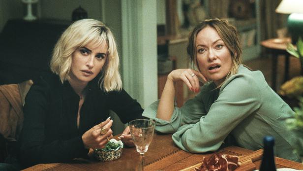 Die Schauspielerinnen Penélope Cruz (li.) und Olivia Wilde an einem Tisch, die eine blickt ernst, die andere überrascht.