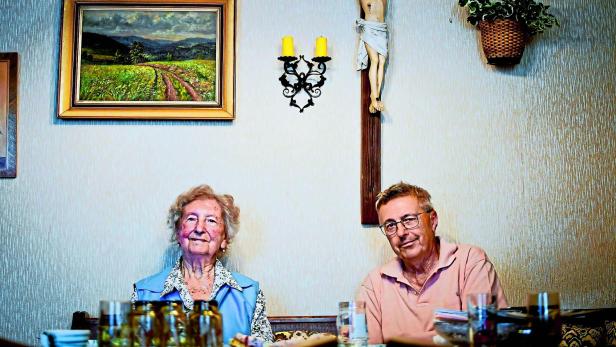 Älteres Paar sitzt am Tisch vor einem Kreuz und einem Landschaftsbild.