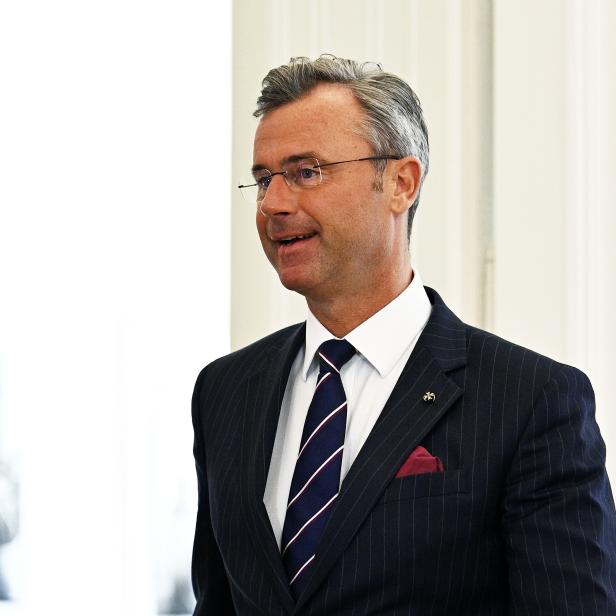 Ein Mann mit Brille trägt einen dunkel gestreiften Anzug, ein weißes Hemd und eine gestreifte Krawatte.
