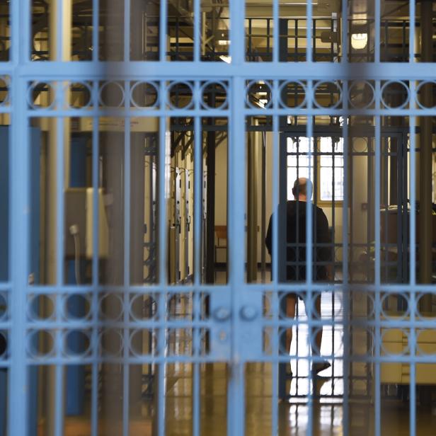Eine Person steht hinter blauen Gitterstäben in einem hellen Flur mit mehreren Türen und Fenstern.