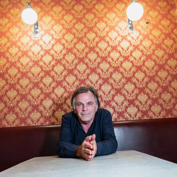 Markus Hinterhäuser sitzt an einem Tisch vor einer Wand mit auffällig orangem Muster.