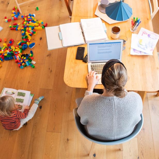 Ein zweijähriges Kind spielt im Wohnzimmer, während seine Mutter im Homeoffice an einem Laptop arbeitet. 