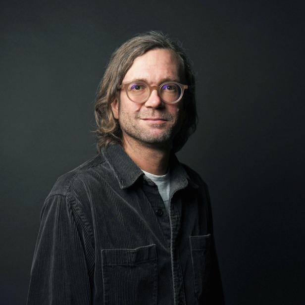 ein Portraitbild von Sebastian Hofer, graues Hemd, grauer Hintergrund