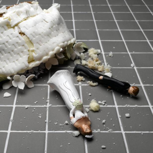 ChatGPT: Auf dem Bild ist ein umgestürztes Stück Hochzeitstorte zu sehen, das auf einem grauen Fliesenboden liegt. Daneben liegen die zerbrochenen Figuren eines Brautpaares, umgeben von Kuchenkrümeln und Zuckerdeko.